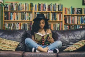 A picture of a young woman of African descent, wearing a fedora and rolled-up jeans, sitting on a couch reading a book with several shelves' worth of books visible behind her. The picture is meant suggest someone deeply engaged with books and reading.