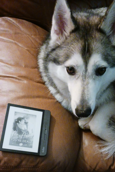 An Alaskan malamute mix with brown eyes looking up at the camera while curled up on a couch. Meanwhile, a Kobo Libra ereader is sitting next to the dog. The ereader is slightly smaller than the dog's head and has a black-and-white e-ink screen.